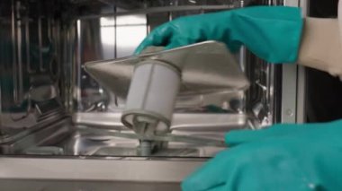 Açık bir bulaşık makinesinin içinde bulaşık makinesi filtresi temizleyen yeşil eldivenli biri. Modern ev aletlerinin bakımı ve hijyeni