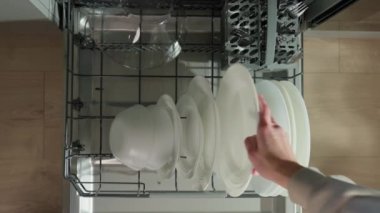 Kadın bulaşık makinesinin alt rafında bulaşık yıkıyor. Kadın çamaşır yıkamak için beyaz tabakları açık bulaşık makinesine yüklüyor. Günlük ev işlerinde modern ev aletleri kullanılıyor.