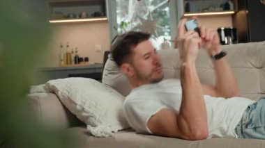 Beyaz tişörtlü ve kot pantolonlu bir adam bej koltukta dinleniyor ve oturma odasında akıllı telefonunu kullanıyor. Erkek kanepede dinleniyor cep telefonuyla sosyal medyada geziniyor