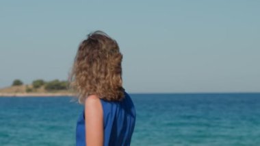 Mavi elbiseli ve güneş gözlüklü rüzgârlı bir günde uçan saçlı bir kadın deniz kıyısında duruyor. Güneşli sahil boyunca yürüyen kadın turist. Yaz seyahati