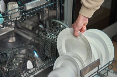 Kadın mutfakta bulaşık makinesinin alt rafından temiz beyaz tabakları boşaltıyor. Modern ev aletlerini kullanarak günlük ev işlerini yapan kişi