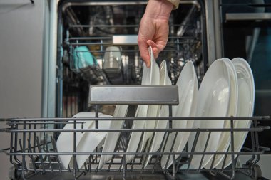 Kadın mutfakta bulaşık makinesinin alt rafından temiz beyaz tabakları boşaltıyor. Modern ev aletlerini kullanarak günlük ev işlerini yapan kişi