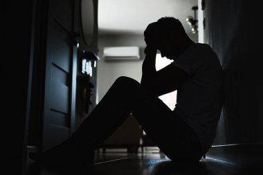 Karanlık odada yerde oturan depresif genç adam. Yalnızlık ve endişeyle kıvranan genç bir erkeğin silueti yüzünü ellerle kapatıyor. Akli dengesi bozuk biri.