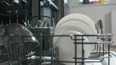 Kadın bulaşık makinesinin alt rafında bulaşık yıkıyor. Kadın çamaşır yıkamak için beyaz tabakları açık bulaşık makinesine yüklüyor. Günlük ev işlerinde modern ev aletleri kullanılıyor.