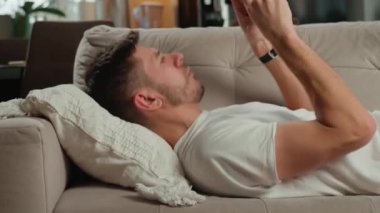 Beyaz tişörtlü ve kot pantolonlu bir adam bej koltukta dinleniyor ve oturma odasında akıllı telefonunu kullanıyor. Erkek kanepede dinleniyor cep telefonuyla sosyal medyada geziniyor