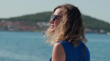 Mavi elbiseli ve güneş gözlüklü kadın yaz tatilinin keyfini çıkarıyor, Hırvatistan 'ın Dalmaçya kıyı şeridinde arka planda manzaralı bir kasabayla birlikte yürüyor.