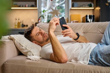 Beyaz tişörtlü ve kot pantolonlu bir adam bej koltukta dinleniyor ve oturma odasında akıllı telefonunu kullanıyor. Erkek kanepede dinleniyor cep telefonuyla sosyal medyada geziniyor