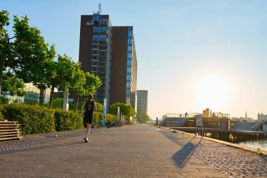 Danimarka, Kopenhag 'da liman boyunca koşan bir sporcu. Gün doğumunda ağaçlarla ve modern binalarla çevrili şehir yolunda koşan bir kadın. Sağlıklı yaşam tarzı kavramı. Kopenhag, Danimarka - 26 Haziran 2024