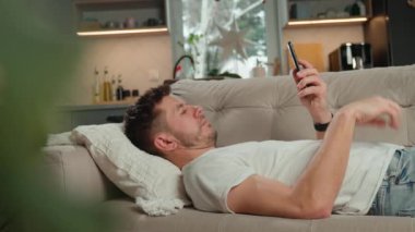 Beyaz tişörtlü ve kot pantolonlu bir adam bej koltukta dinleniyor ve oturma odasında akıllı telefonunu kullanıyor. Erkek kanepede dinleniyor cep telefonuyla sosyal medyada geziniyor