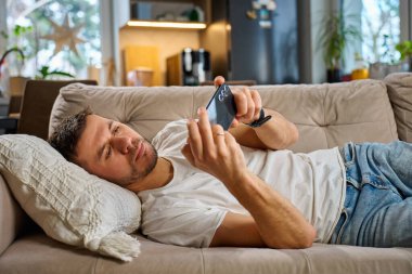 Beyaz tişörtlü ve kot pantolonlu bir adam bej koltukta dinleniyor ve oturma odasında akıllı telefonunu kullanıyor. Erkek kanepede dinleniyor cep telefonuyla sosyal medyada geziniyor