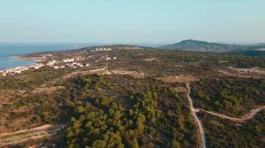 Yeşil tepelerle çevrili kıyı köyünün havadan görünüşü, virajlı toprak yollar ve arka planda deniz. Doğal Akdeniz manzarası