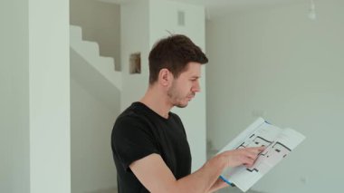 Siyah gömlekli bir adam parlak iç mekanına bakıyor ve tamamlanmamış bir dairede mimari kat planını inceliyor. Gayrimenkul yatırımı ve yenileme planı kavramı