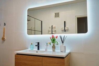 Ahşap lavabo dolabı, siyah musluğu ve dekoratif unsurları olan şık bir banyo. Modern apartmandaki aydınlatmalı büyük LED aynalı modern banyo tasarımı.
