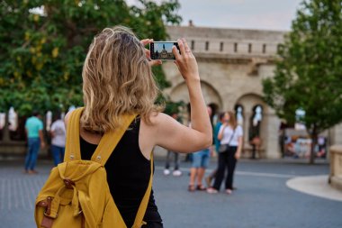 Sarı sırt çantalı kadın turist seyahat sırasında Budapeşte 'de tarihi eserlerin fotoğrafını çekiyor. Avrupa 'da gezmek ve gezmek için akıllı telefon kullanan bir kadın