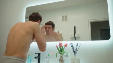 Gömleksiz bir adam banyo aynasında yansımasına bakıyor, yüzünü kontrol ediyor, yüz kırışıklığı için endişeleniyor. Yaşlanma önleyici, cilt bakımı ve kişisel bakım konsepti. Sabah hijyeni.