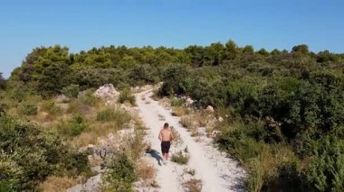 Gömleksiz bir adam sıcak güneşli bir günde toprak yolda koşuyor, yemyeşil doğa ve hava manzarası ile çevrili. Profesyonel sporcu dayanıklılığı eğitimi. İz çalıştırma ve uygunluk kavramı