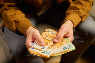 Evdeki Euro banknotlarını sayan adama yakın çekim. Kişisel mali durumunu planlayan, nakit parayla koltukta oturan biri. Ev bütçesi, tasarruf ve kişisel finans planlama kavramı