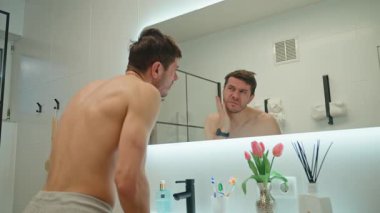 Gömleksiz adam banyo aynasında kendine bakıyor. Kırışıklıklarını ve saçlarını kontrol eden bir adam. Sabah hijyeni.