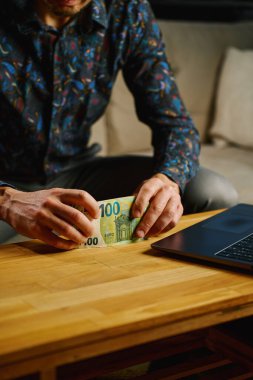 Elinde 100 avroluk banknotlar olan bir adam masanın önünde dizüstü bilgisayarla oturuyor. Evde nakit para sayan biri var. Kişisel finans ve ev bütçesi kavramı