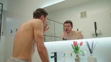 Gömleksiz bir adam banyo aynasında yansımasına bakıyor, yüzünü kontrol ediyor, yüz kırışıklığı için endişeleniyor. Yaşlanma önleyici, cilt bakımı ve kişisel bakım konsepti. Sabah hijyeni.