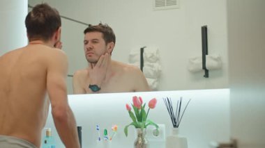 Aydınlanmış banyo aynasının önünde yüzü ve derisini kontrol eden gömleksiz bir adam. Sabah temizliği ve kişisel bakım.