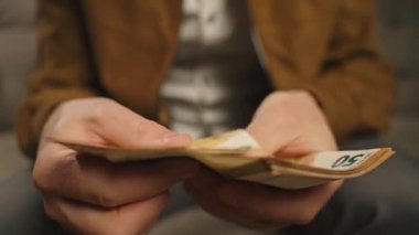 Evdeki Euro banknotlarını sayan adama yakın çekim. Kişisel mali durumunu planlayan, nakit parayla koltukta oturan biri. Ev bütçesi, tasarruf ve kişisel finans planlama kavramı