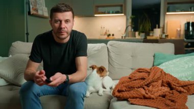 Kanepede oturan ve elinde uzaktan kumanda tutan bir adam. Evdeki kanepede evcil köpekle televizyon izleyen bir erkek. Rahatlama, yaşam tarzı ve ev hayatı kavramı