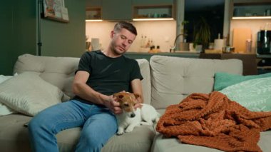 Adam kanepede oturuyor, köpeğiyle oynuyor. Oturma odasında kanepede köpek besleyen sıradan bir adam, evcil hayvanla eğleniyor. Rahatlama ve evcil hayvan bakımı kavramı.