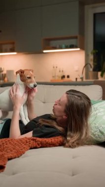 Kanepede uzanmış köpeğiyle oynayan bir kadın. Evdeki kanepede Jack Russell Terrier köpeğiyle bağ kuran mutlu kadın. Şefkat, rahatlama ve evcil hayvan sevgisi kavramı