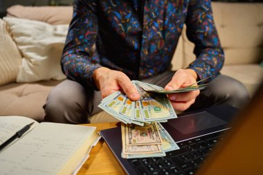 Elinde dolar banknotları olan bir adam, masanın önünde dizüstü bilgisayar ve defterle oturuyor. Faturaları ve giderleri ödedikten sonra kalan parayı sayan kişi. Kişisel finans ve ev bütçesi