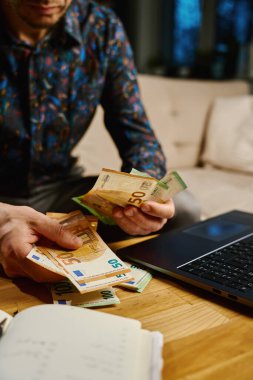 Avro banknotları tutan, dizüstü bilgisayarı ve defteriyle masanın önünde oturan bir adam. Faturaları ve giderleri ödedikten sonra kalan parayı sayan kişi. Kişisel finans ve ev bütçesi