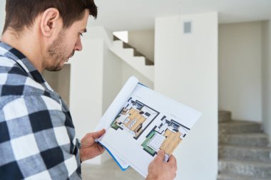 Düşünceli bir adam bitmemiş bir evde dururken kat planını inceliyor. Erkek ev sahibi, yeni apartmanın mimari planlarını inceliyor. İç tasarım ve yenileme kararları kavramı