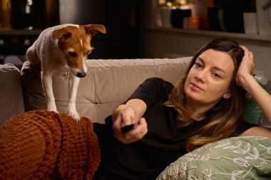 Kanepede uzanmış elinde uzaktan kumanda olan bir kadın. Kanepede evcil köpeğiyle televizyon izleyen bir kadın. Rahatlama, yaşam tarzı ve ev hayatı kavramı