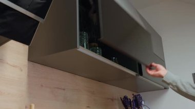 Adam üst mutfak dolabını açar ve içki bardağını alır. El, dolabı şeffaf bardaklarla açar.