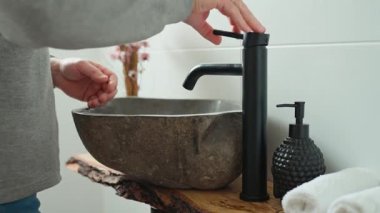 Adam ellerini taştan bir lavaboda siyah modern muslukla yıkıyor. Banyo içinde akan suyun altında taş havzayla yakın plan çekilmiş.