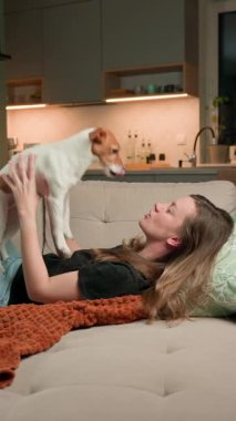Kanepede uzanmış köpeğiyle oynayan bir kadın. Evdeki kanepede Jack Russell Terrier köpeğiyle bağ kuran mutlu kadın. Şefkat, rahatlama ve evcil hayvan sevgisi kavramı