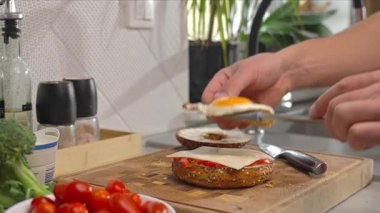 Kırmızı gömlekli adam kızarmış yumurta, peynir ve jambon dilimlerini simit yarısına koyuyor. Kahvaltı sırasında erkeklerin ellerinin sandviçi hazırladığı yakın plan. Evde sağlıklı bir sabah yemeği hazırlığı.