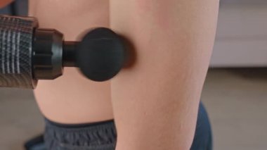 Atletik adam içeride perküsyon masaj aleti kullanıyor. Adam oturma odasında otururken kol kaslarına masaj tabancası uyguluyor. Egzersiz sonrası tedavi görüyor. Ev zindelik ve rahatlama kavramı