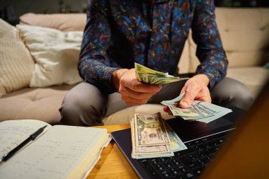 Elinde dolar banknotları olan bir adam, masanın önünde dizüstü bilgisayar ve defterle oturuyor. Faturaları ve giderleri ödedikten sonra kalan parayı sayan kişi. Kişisel finans ve ev bütçesi