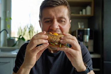 Adam yumurtalı, peynirli, domatesli ve yeşillikli ekmeklerden büyük bir ısırık alıyor. Aç adam mutfakta sandviç yiyor, kapat.