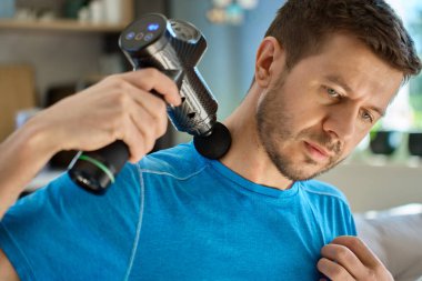 Adam boyun ve omuz bölgesindeki gerilimi azaltmak için masaj tabancası kullanıyor. Erkek sporcu antrenmandan sonra iyileşmek için kendine masaj yapıyor.