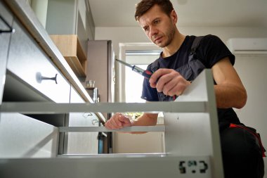 Adam modern dolapta tornavida kullanarak mutfak çekmecesini monte ediyor. Tamirci, çağdaş mutfağa çekmece kaydırma mekanizması kuruyor ve ayarlıyor. Mobilya kurulumu