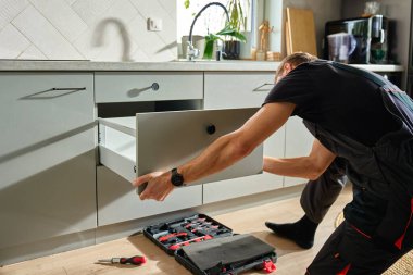 Adam modern dolapta tornavida kullanarak mutfak çekmecesini monte ediyor. Tamirci, çağdaş mutfağa çekmece kaydırma mekanizması kuruyor ve ayarlıyor. Mobilya kurulumu