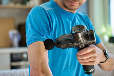 Atletik adam içeride perküsyon masaj aleti kullanıyor. Adam oturma odasında otururken kol kaslarına masaj tabancası uyguluyor. Egzersiz sonrası tedavi görüyor. Ev zindelik ve rahatlama kavramı