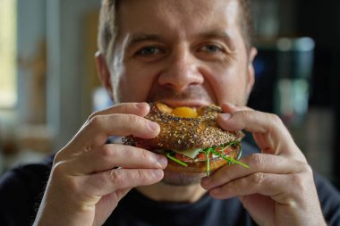 Adam yumurtalı, peynirli, domatesli ve yeşillikli ekmeklerden büyük bir ısırık alıyor. Aç adam mutfakta sandviç yiyor, kapat.