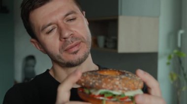 Adam yumurtalı, peynirli, domatesli ve yeşillikli ekmeklerden büyük bir ısırık alıyor. Aç adam mutfakta sandviç yiyor, kapat.