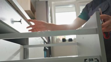 Adam modern dolapta tornavida kullanarak mutfak çekmecesini monte ediyor. Tamirci, çağdaş mutfağa çekmece kaydırma mekanizması kuruyor ve ayarlıyor. Mobilya kurulumu