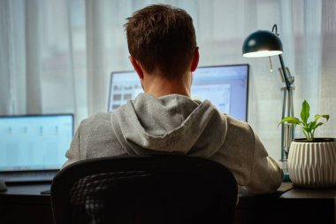 Gri kapüşonlu adam bilgisayar masasında çift monitörlü ve arka planda lambayla çalışıyor. Dahili ofiste uzaktan çalışan serbest çalışan biri. Uzak çalışma ve üretkenlik kavramı