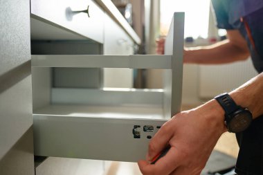 Adam modern dolapta tornavida kullanarak mutfak çekmecesini monte ediyor. Tamirci, çağdaş mutfağa çekmece kaydırma mekanizması kuruyor ve ayarlıyor. Mobilya kurulumu