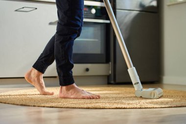 Yalınayak adam mutfaktaki ahşap zeminde örülmüş halıyı süpürüyor. Dairesinde el yapımı elektrikli süpürge kullanıyor. Günlük ev temizliği ve ev işleri kavramı